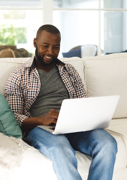 Smiling african american man using laptop and sitting on couch in living room. spending time alone at home with technology.